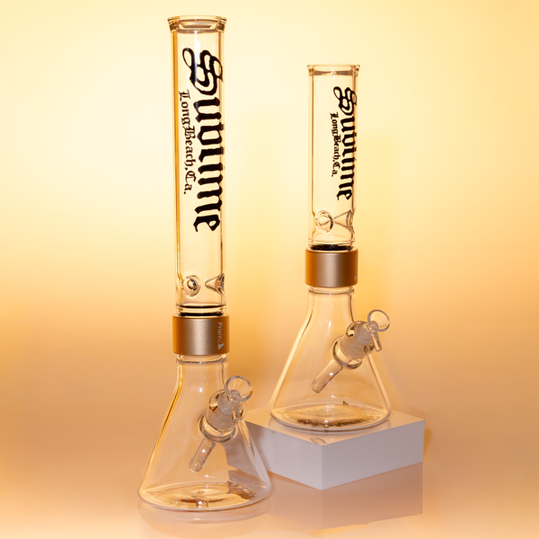 Size comparison: Prism x Sublime Official Clear Beaker collection. Featuring the "Tall" vs "Standard" modular bong heights. Both models showcase the Opie Ortiz sun logo on the beaker base and the Sublime Long Beach gothic branding. This shot helps collectors visualize the scale and volume difference of these official band-collaboration glass pieces.